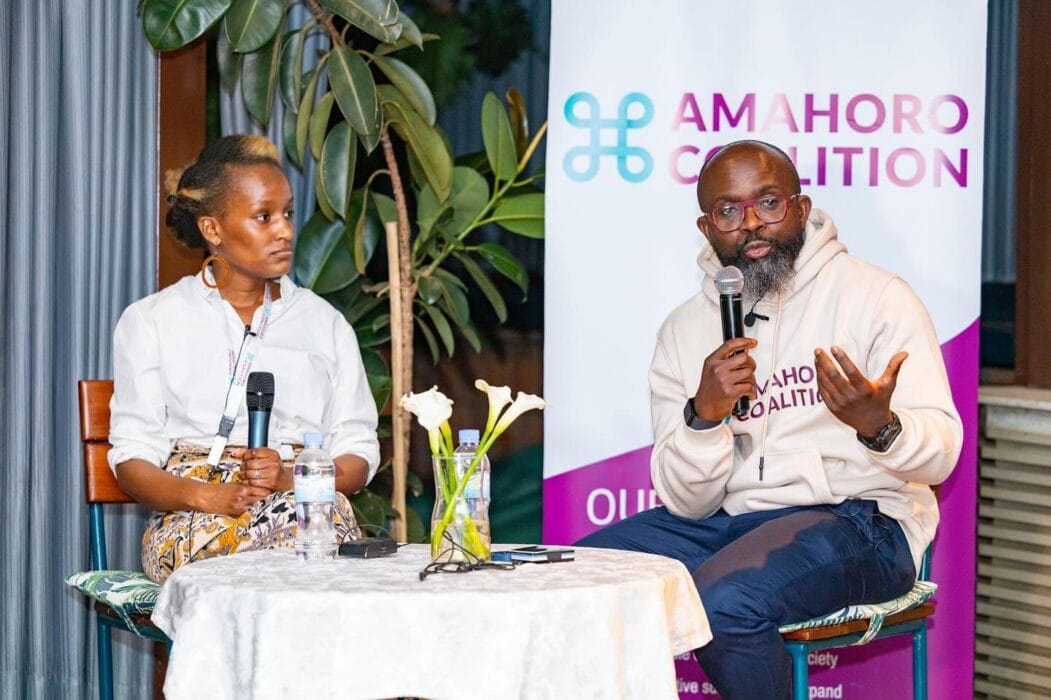Smiling woman and man participating in an AMAHORO Coalition event, engaging in a discussion while holding microphones, surrounded by a lush indoor setting with plants and a SMEPEAKS banner, promoting community collaboration.