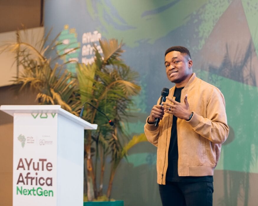 Youth African entrepreneur giving a presentation at AEYE Africa NextGen event with a microphone, standing by a podium with event branding, surrounded by tropical plants and a vibrant green themed backdrop.