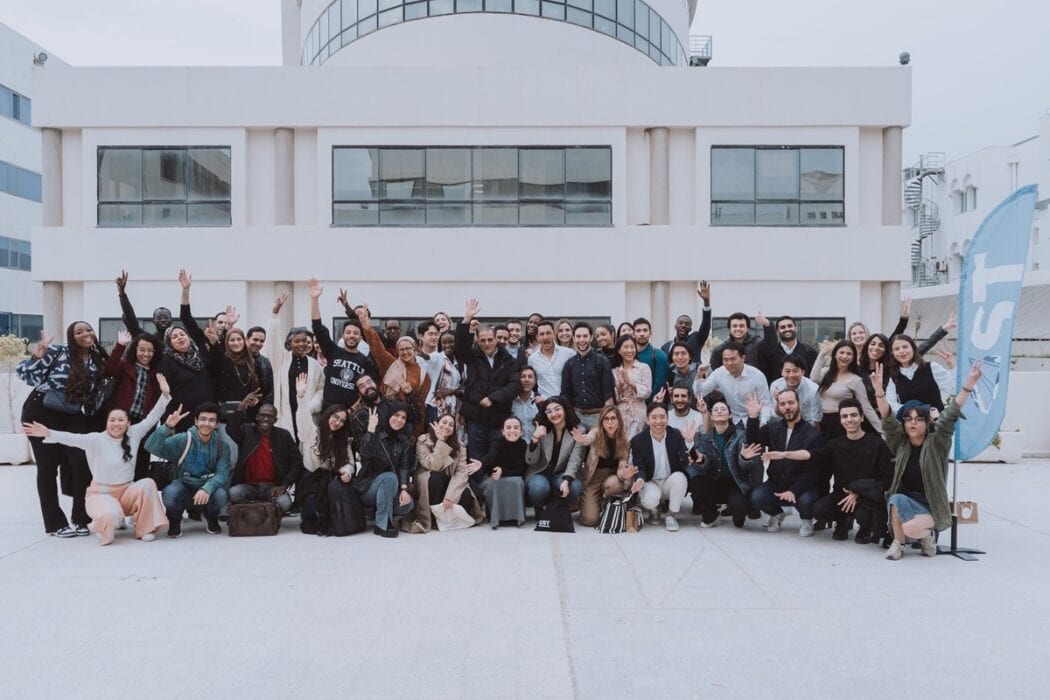 Large diverse group of people smiling and waving in front of a modern building during a corporate event or team-building activity.